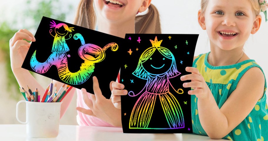 two girls holding scratch artwork up at table