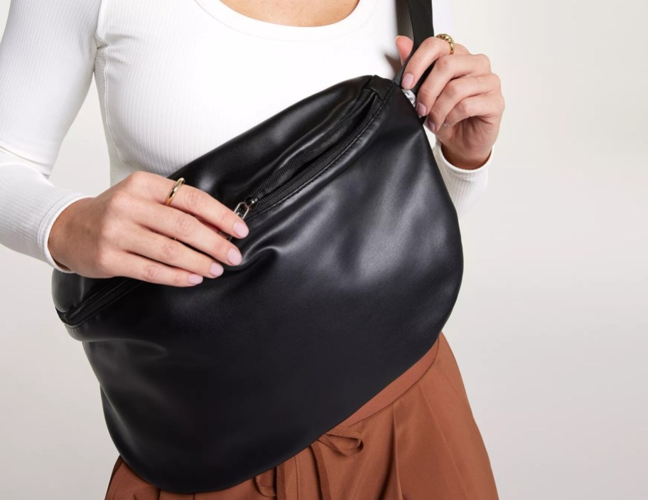 black faux leather sling bag over front of womans body with one hand holding top of bag and other hand holding strap