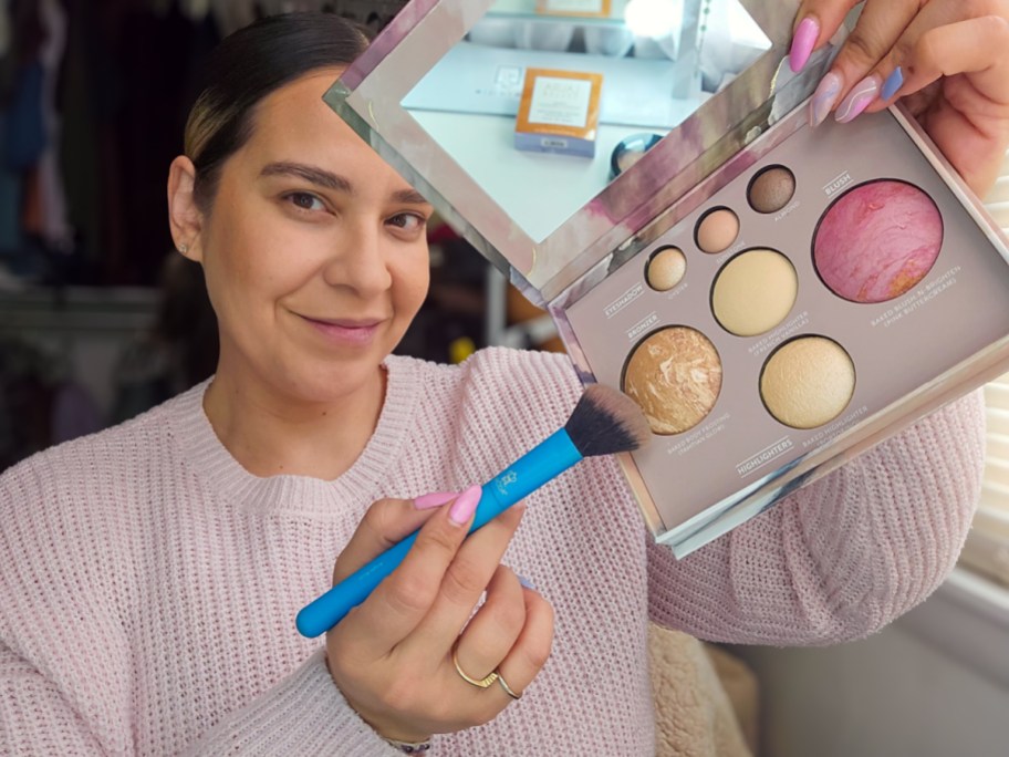 smiling woman tapping a makeup brush to a laura geller makeup palette held aloft