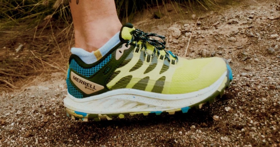 woman's foot wearing a yellow, teal, black and white Merrell trail shoe while walking on a trail