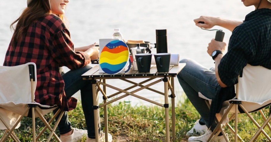 people camping sitting in chairs at a table with drinks and a colorful bluetooth speaker playing music