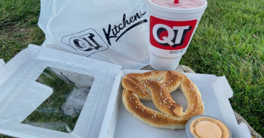 Pretzel with cup of cheese in an open cardboard box with a soda next to it. All are sitting in the green grass.