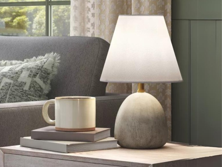 greyish wood color lamp with white shade sitting on a side table next to a large coffee mug and books