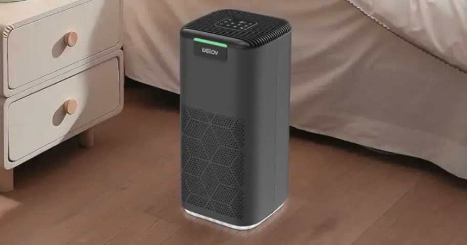 grey air purifier in a bedroom next to a nightstand and bed
