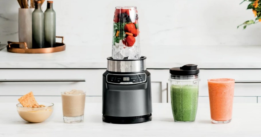 Ninja blender with smoothie cup on it, 1 beside it and other glasses and bowls of blended items on a countertop