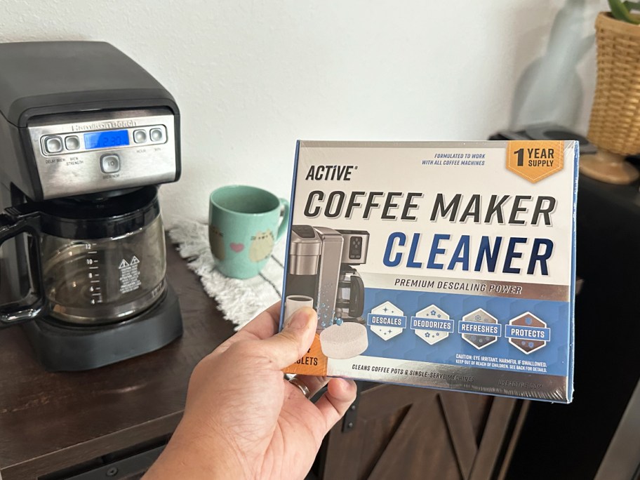 hand holding a box of Active Coffee Maker Cleaner near coffee maker