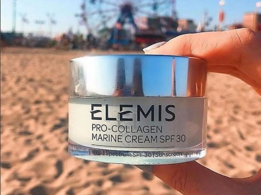 A hand holding an Elemis cream on a beach