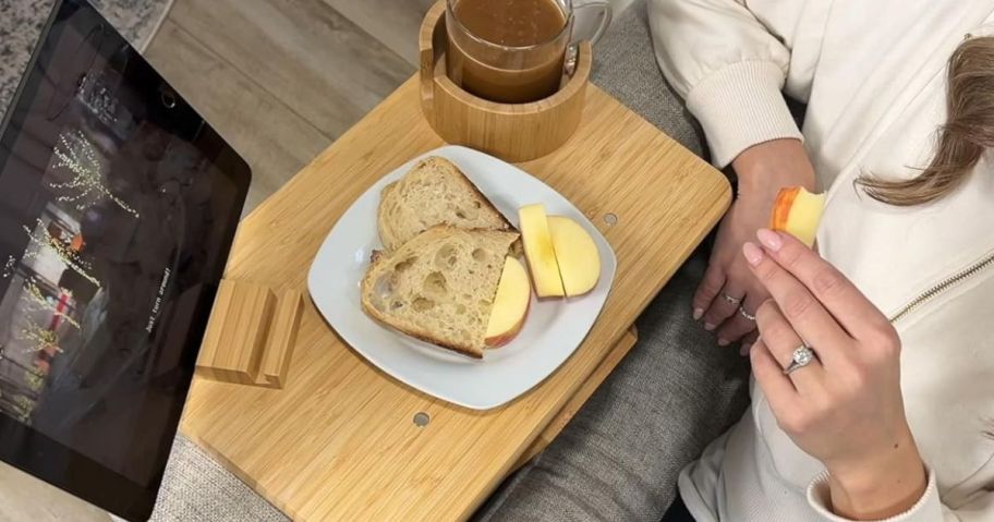 A woman watching a tablet with a bamboo tray attached to their couch 