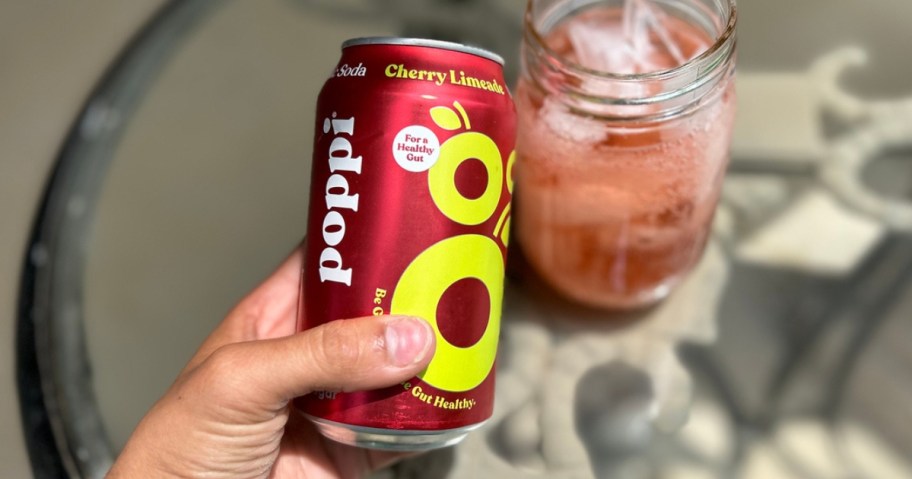 hand holding a Cherry Lime Poppi Soda can with a glass of soda behind it