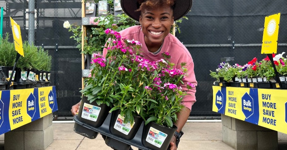 woman smiling at the camera while holding 3 flowers in planters in the garden center at Lowe's