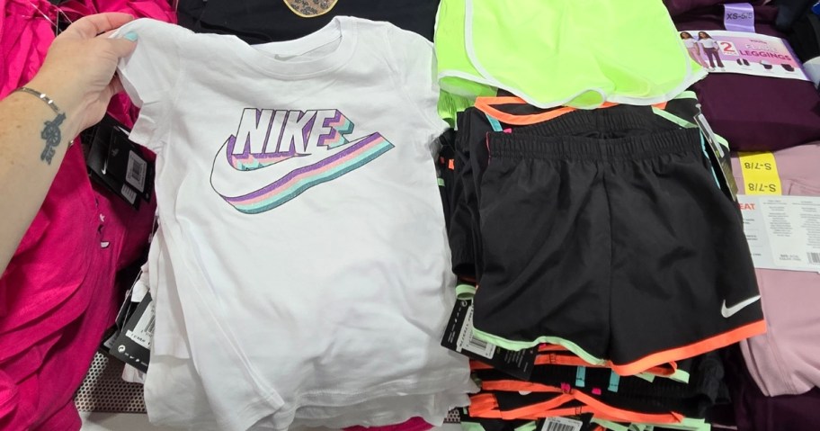 hand reaching for a kid's Nike logo tshirt in white next to Nike kid's shorts in black with neon trim