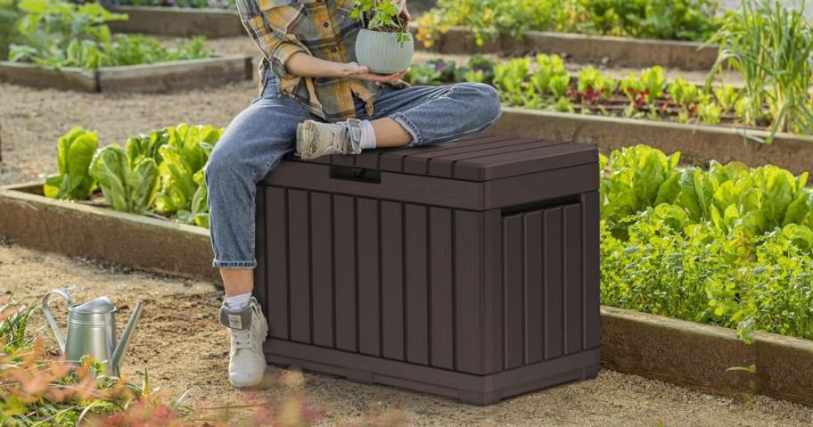A woman sitting on a Keter Kentwood 50 Gallon Resin Deck Box near a garden