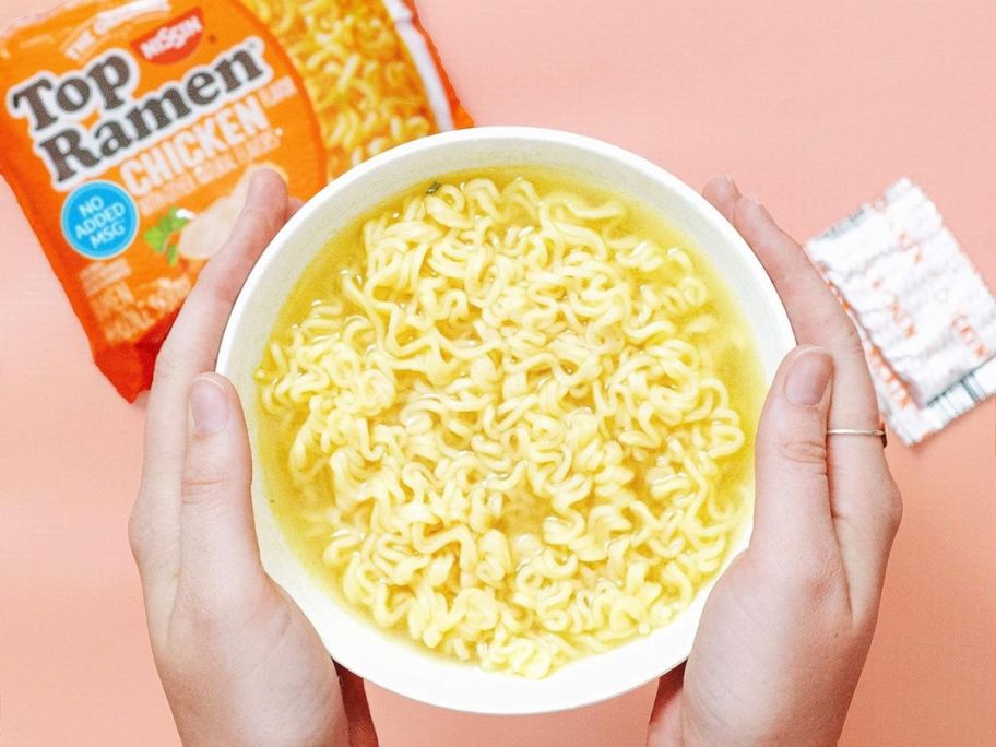 a pair of hands holding a bowl of prepared ramen noodle soup