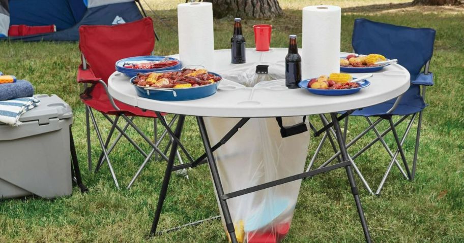 An Ozark Trail Camping table with food and drinks set on it and a trash bag hanging in the center