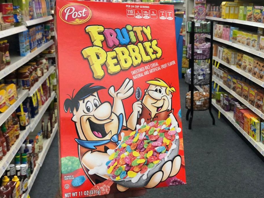 Hand holding up a box of Fruity Pebbles Cereal in the middle of an aisle at the store