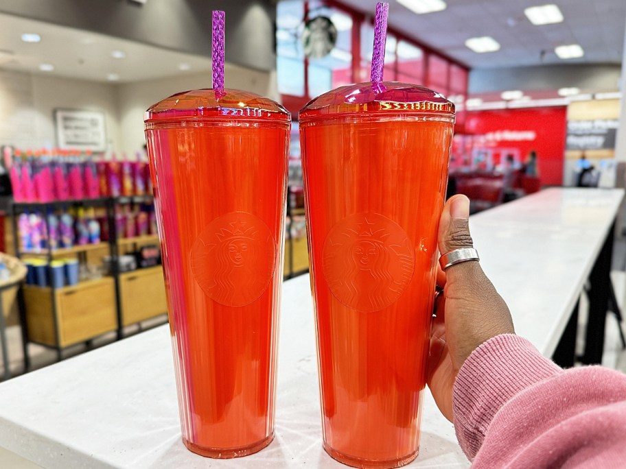 hand touching two red starbucks cold cups with dome lids and pink straws