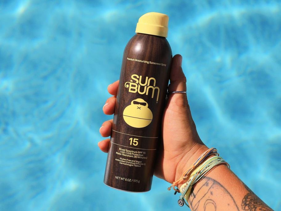 A hand holding a Sun Bum Original SPF 15 Sunscreen Spray over a pool