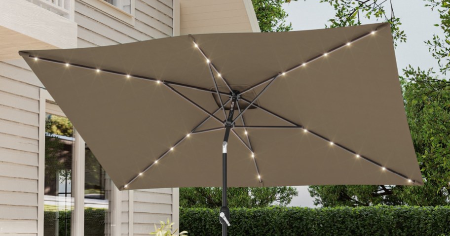 A tan 10-foot tilt market patio umbrella featuring built-in string lights along its ribs, set up outdoors on a patio next to a house.