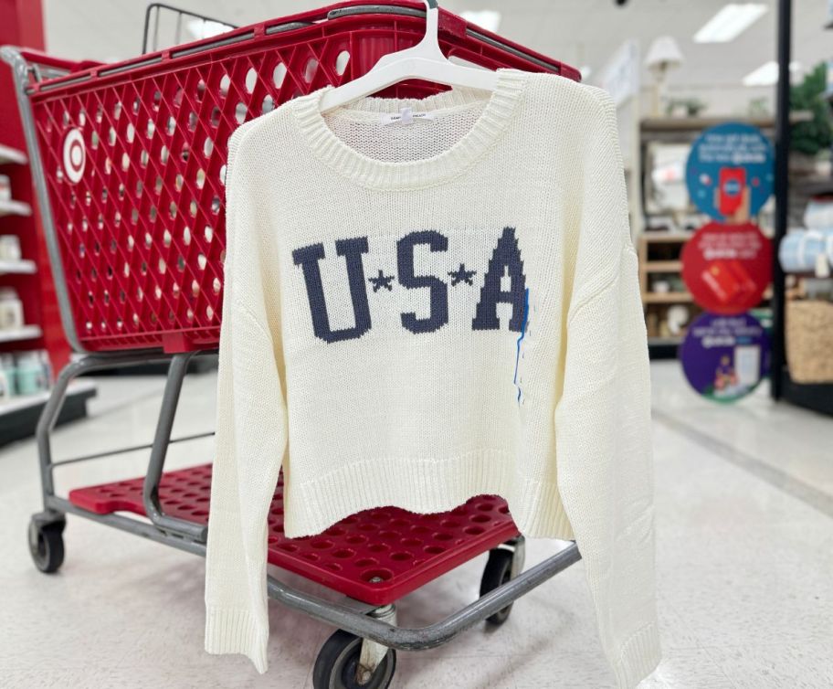 a womens white USA sweater hanging on the side of a shopping cart in a target store