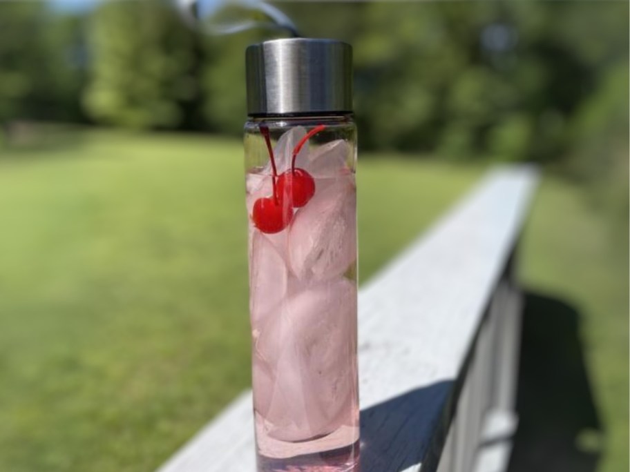 clear water bottle with a sliver lid and carry strap with ice and a pink drink and cherries sitting on a deck rail