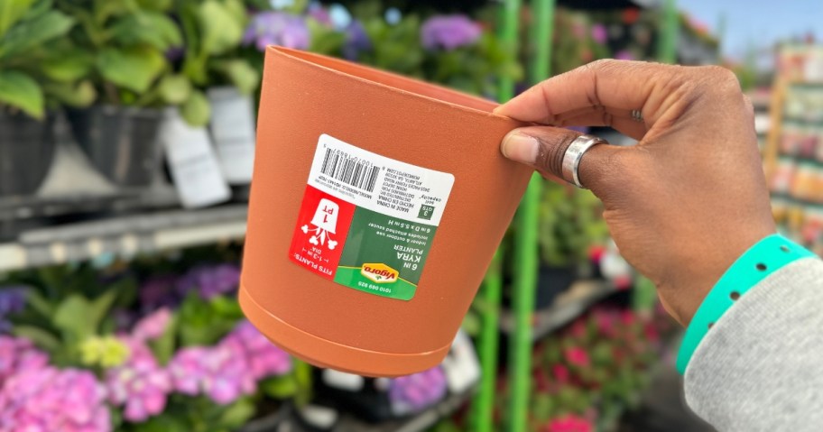 hand holding a terracotta colored small plastic planter, flowers in the background in a garden center