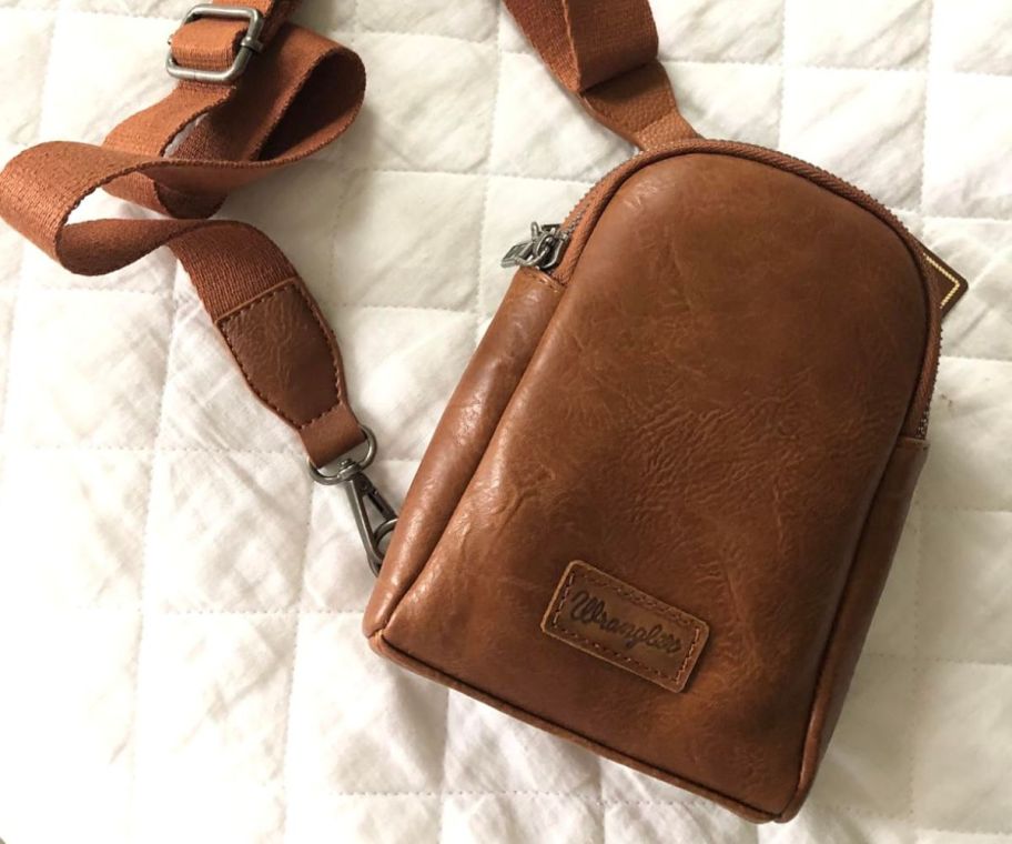 A Wrangler Crossbody Bag in Brown on a bed