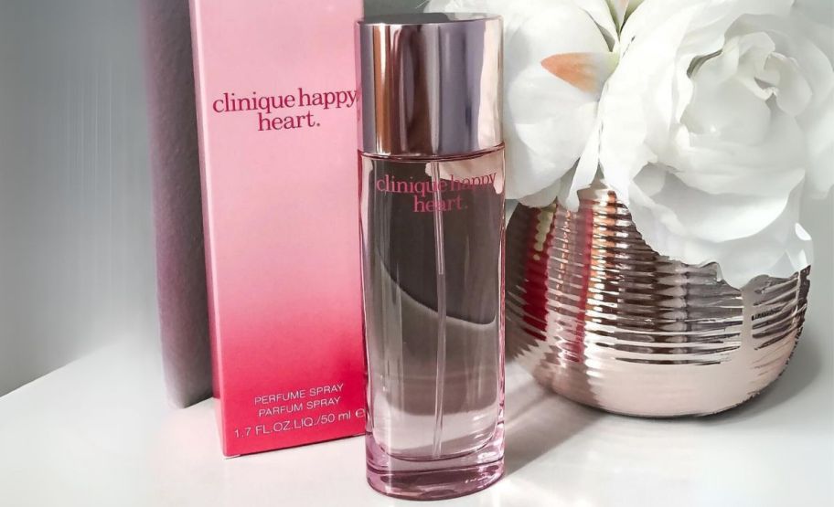 a bottle of clinique happy heart perfume with packaging sitting next to a white flower in a silver vase