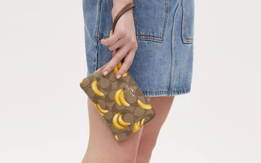 a womans hand holding a coach banana print corner zip wristlet