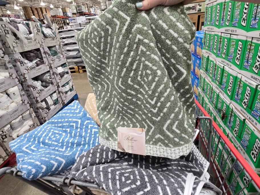 hand holding a bath towel with a geometric print in green, more colors in a shopping cart behind it