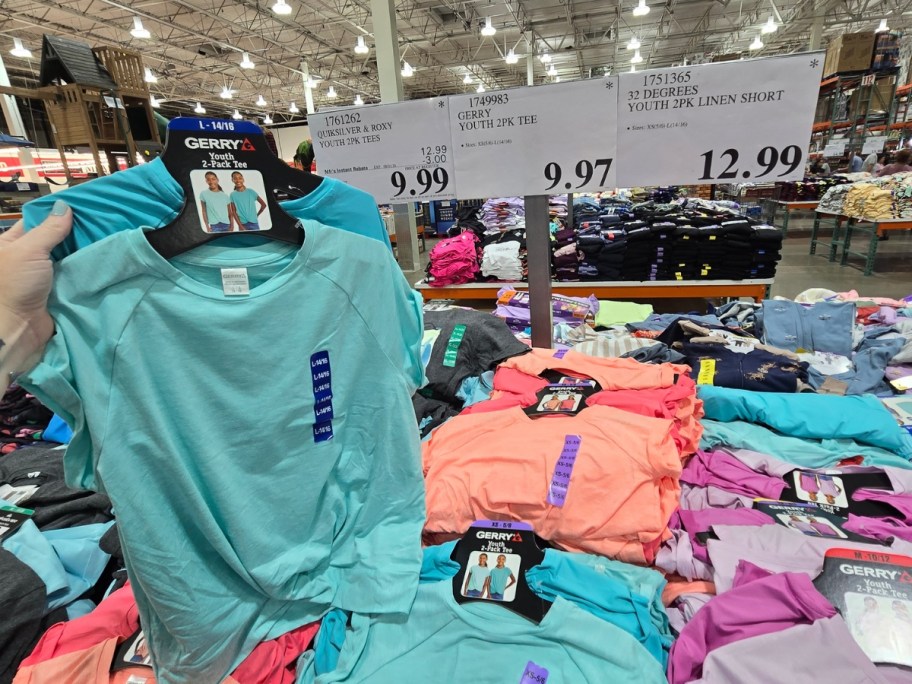 hand holding a 2 pack of kids teshirts in a blue green color with more on a display behind it