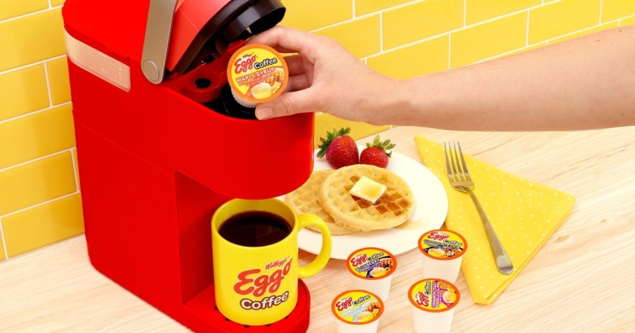 person putting an Eggo coffee k-cup pod into a red Keurig machine, yellow Eggo Coffee mug in the machine, plate of waffles and more k-cups to the side