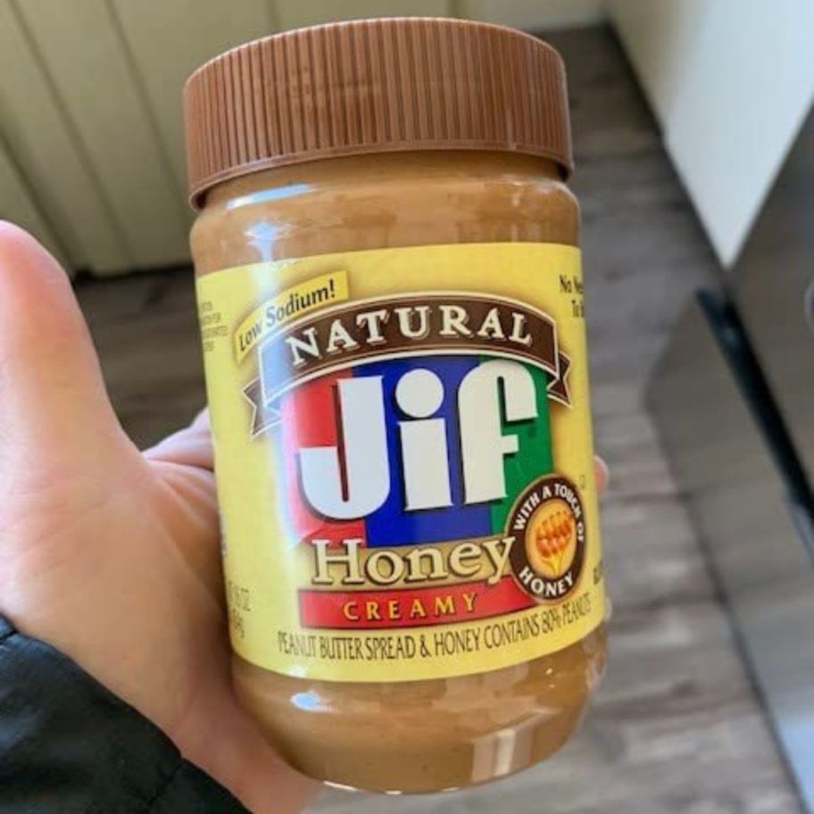 a man's hand holding a jar of jif peanut butter with honey