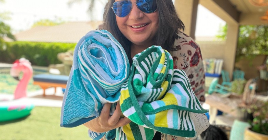 Lina outside holding different style Kohl's beach towels that are rolled up, large flamingo float and swimming pool behind her
