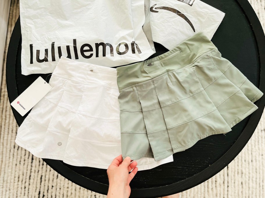 hand holding white lululemon skirt and green amazon skirt with bags behind them