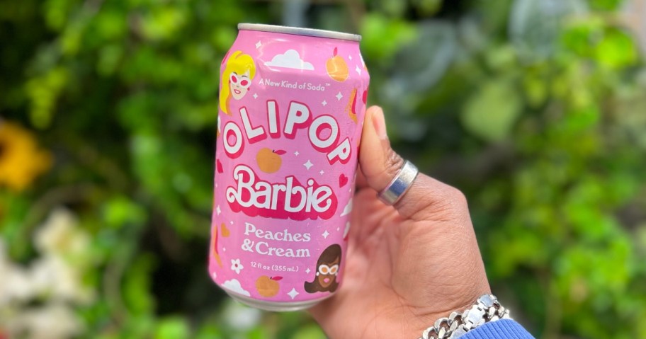 hand holding a pink color can of Olipop Barbie soda alternatives, with greenery in the background