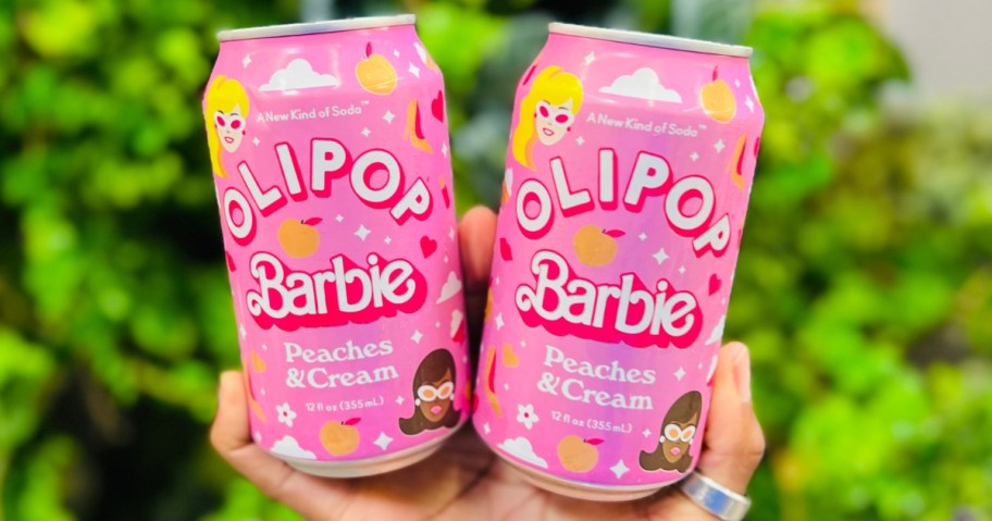 hand holding 2 pink color cans of Olipop Barbie soda alternatives, with greenery in the background