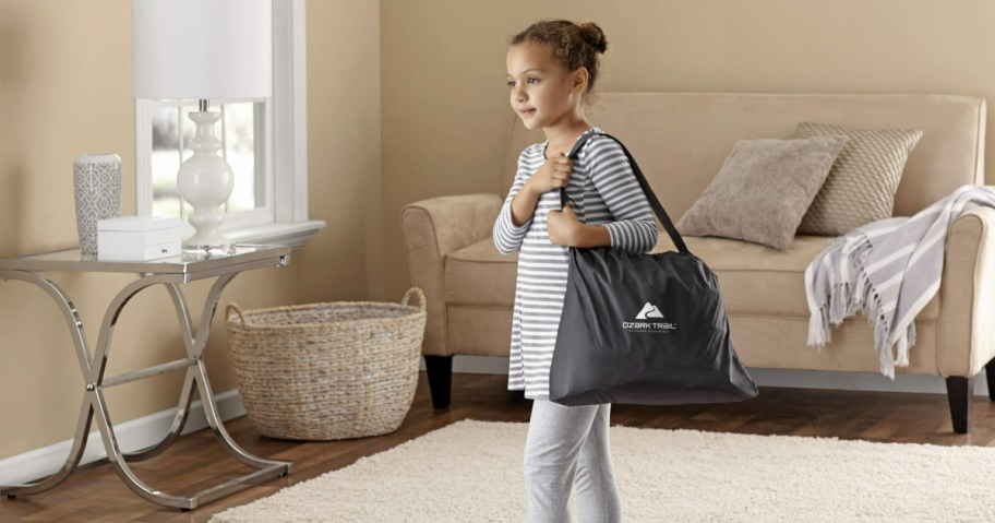 little girl in a living room carrying an ozark trail air mattress bag