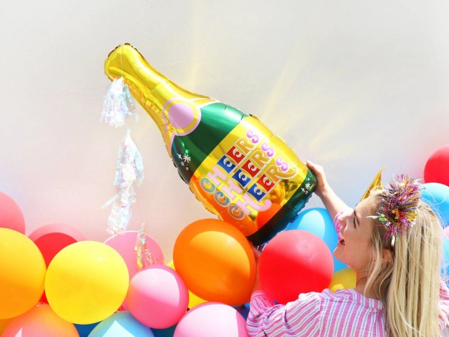 woman putting a large champagne bottle balloon up on a wall above a balloon banner