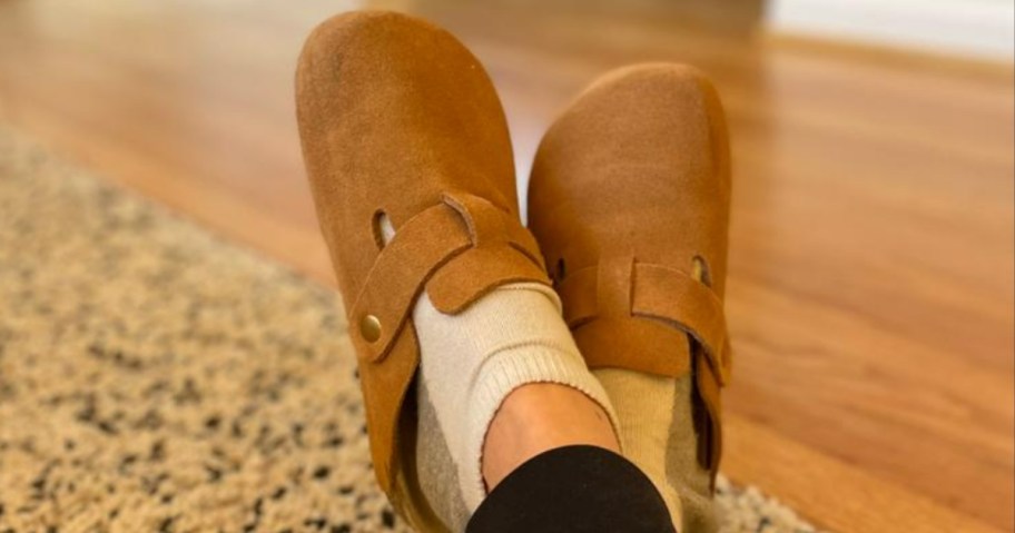 feet propped up and crossed wearing a brown color of suede clog mules with off white socks