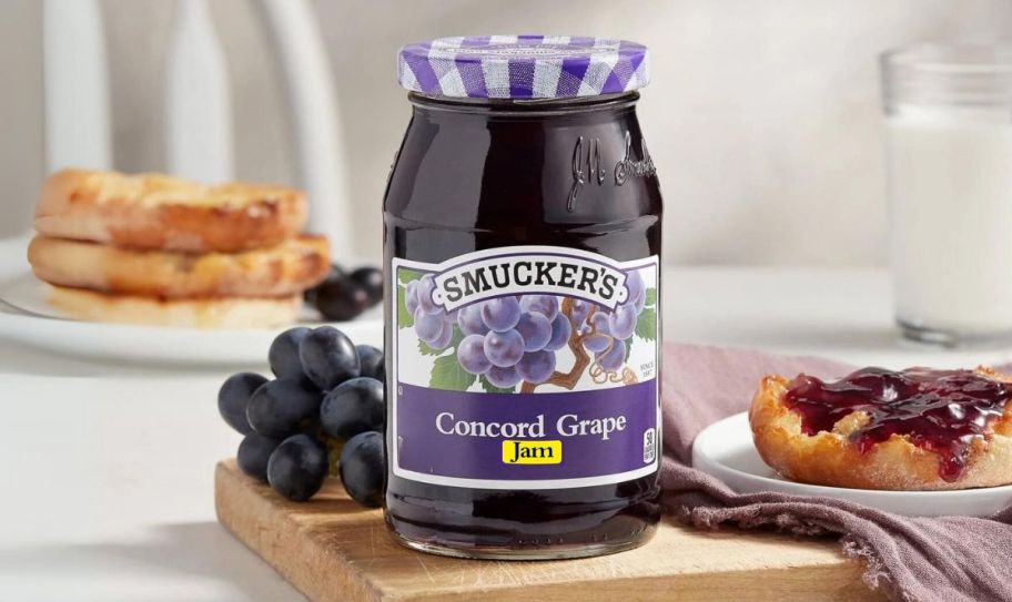 a jar of grape jam on a table shown with english muffins and fresh grapes