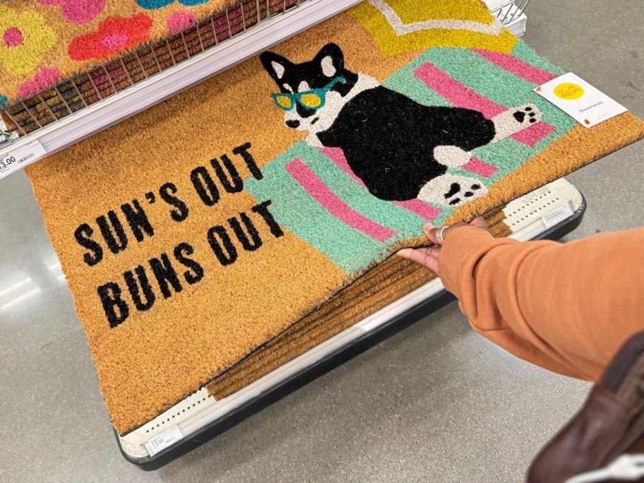hand reaching for a coir doormat that has a black Corgi dog laying on a beach towel that says "sun's out, buns out"...