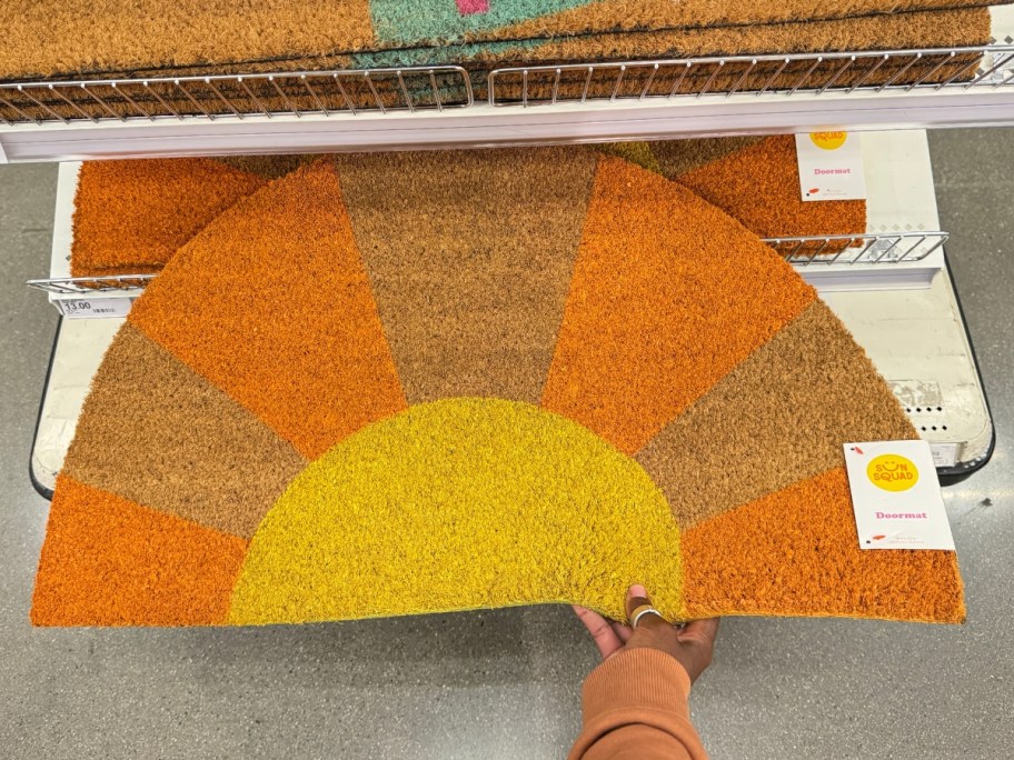 hand pulling a half circle coir doormat with yellow and orange sun rays out of a display bin