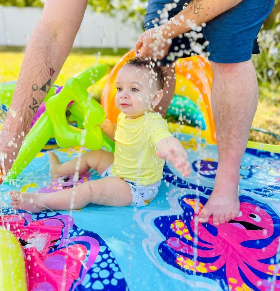 baby sitting on colorful splash pad with man standing in grass