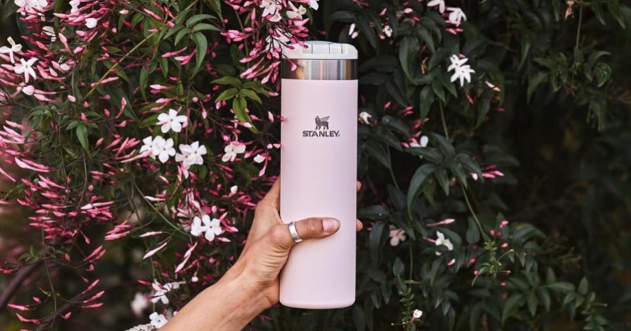 woman's hand holding a light pink flip top Stanley water bottle, pink and white flowers behind it