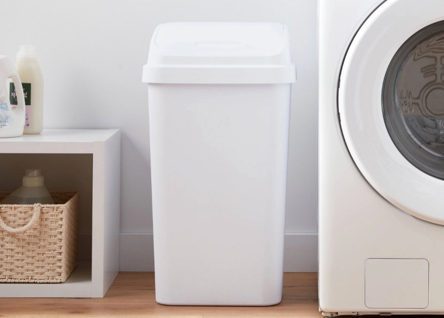 a white plastic flip top trash can next to a washing machine