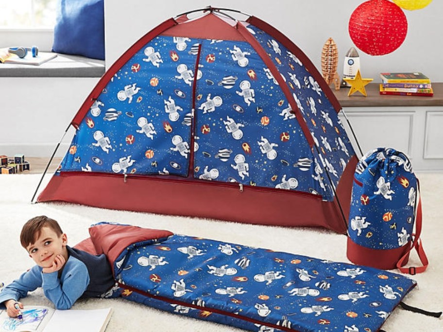 little boy in a blue sleeping bag with space images with a matching tent behind him and a matching carry bag