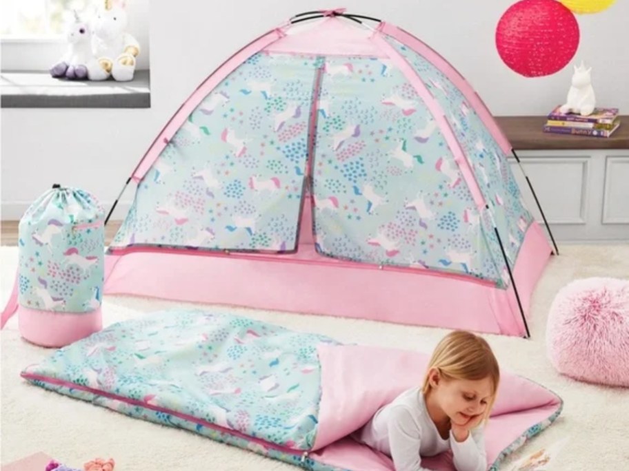 little girl in a pink and blue sleeping bag with unicorns on it with a matching tent behind him and a matching carry bag