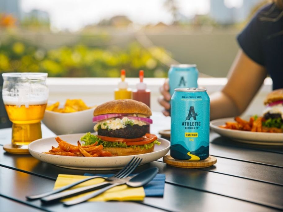 A table with burgers on plates and cans and a glass of Athletic Brewing Co non-alcoholic beer