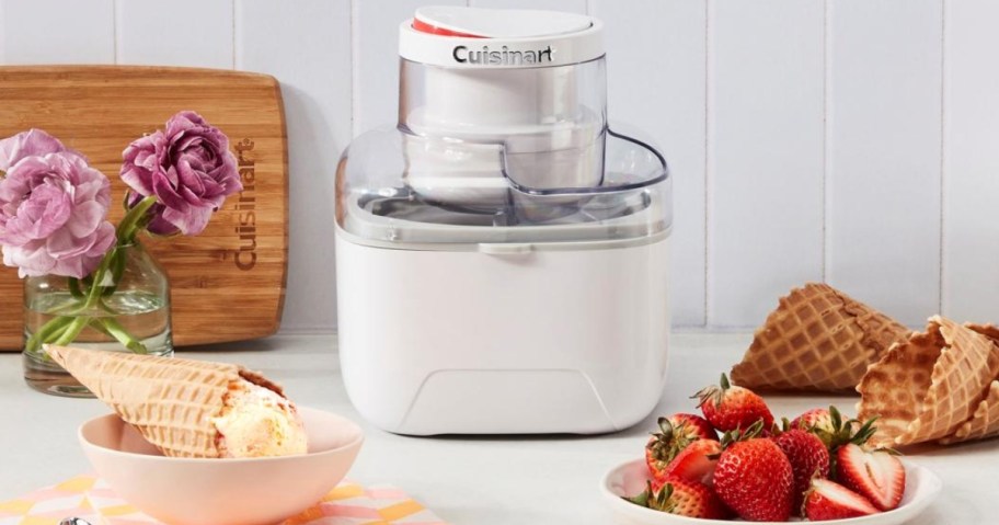 small white Cuisinart Ice Cream Maker on a counter with fruit on 1 side, and a waffle cone with ice cream in a bowl on the other side