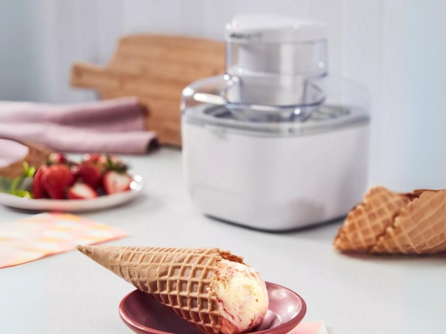 small white Cuisinart Ice Cream Maker on a counter with fruit and a waffle cone with ice cream in a bowl